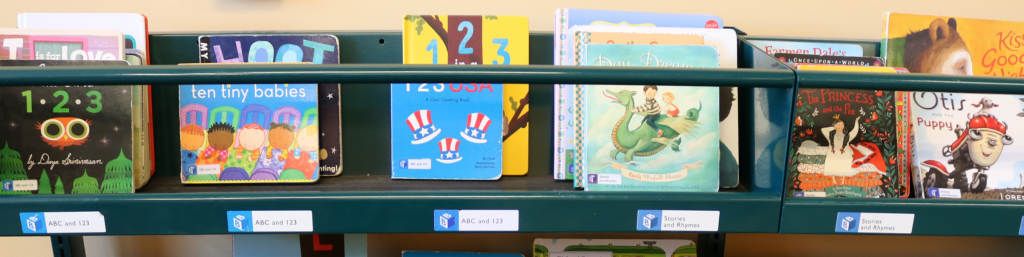 A shelf of board books in a library, with labeled stickers along the bottom of the shelves denoting the subject of the books.